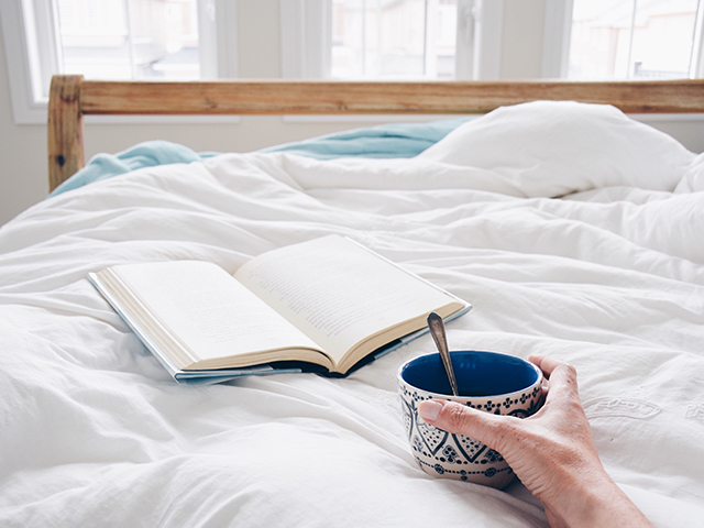 Book laying on bed and woman holding a coffee cup.
