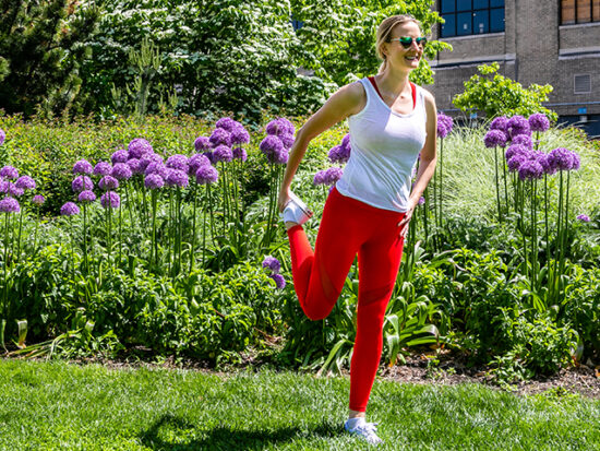 Megan stretching on the grass in front of flowers wearing red Fabletics leggings.